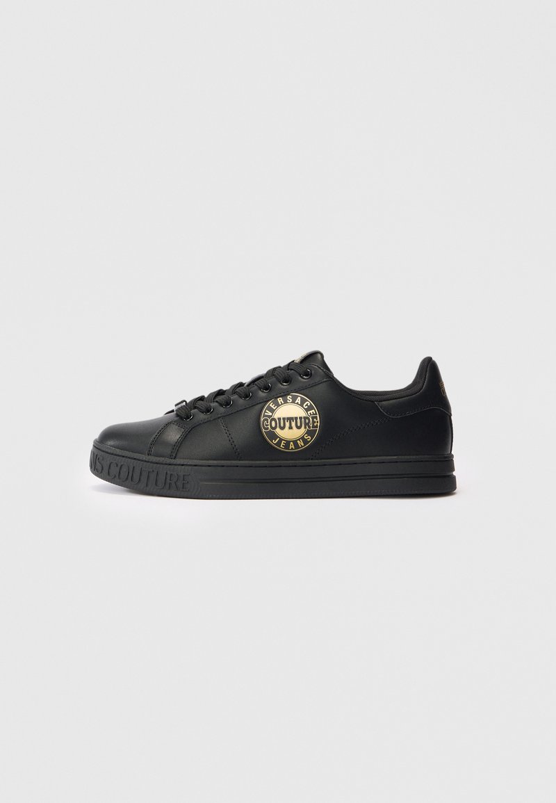 Črne nizke teniske z zlatim logotipom "Versace Jeans Couture" na strani in vtisnjeno blagovno znamko na podplatu, prikazane v profilu na beli podlagi.