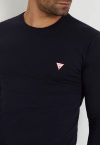 Svart långärmad T-shirt i slätt tyg med en röd och vit triangel-logotyp på vänster bröst. Tätt sittande design, rundad halsringning.