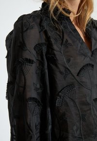 Blusa nera trasparente con intricati ricami floreali, dettagli strutturati e una vestibilità rilassata con colletto.