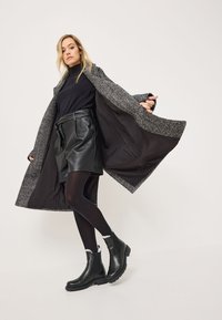 Dlouhý černý kabát s šedým vzorovaným límcem, vrstvený přes černý žebrovaný top a koženou sukni. Modelka má na sobě černé vysoké boty po kolena.