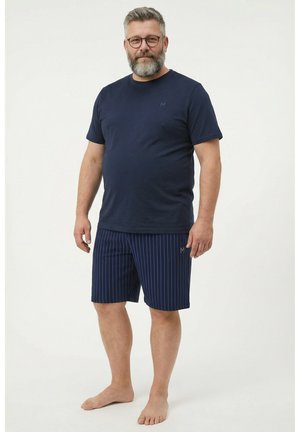 Skægget mand iført briller med navyblå t-shirt og navyblå stribede shorts, stående barfodet på hvid baggrund.