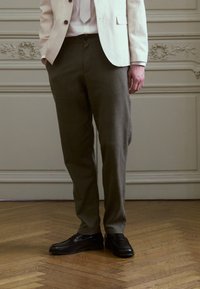 Light-colored linen blazer over a white shirt and dark green trousers. Black leather shoes complete the outfit. Wood floor background.