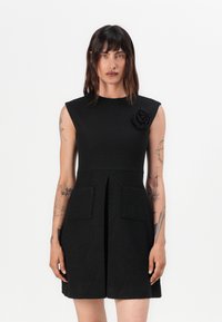 Black sleeveless dress with textured fabric, a fitted bodice, pleated skirt, and two front pockets. Features a flower detail on the shoulder.