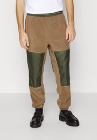 Brown fleece pants with green nylon panels, tapered leg design, elastic waistband, and side pockets. Textured fabric with contrasting colors.