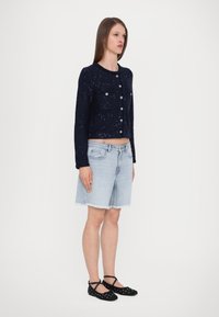 Marine teksturert cropped jakke med knappeåpning og to brystlommer, sammen med lyseblå denimshorts og svarte prikkete ballerinas.