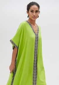 Limegrön kaftan med V-ringning, korta ärmar och en kontrasterande svart och vit mönstrad kant längs kanterna. Lättviktsmaterial.