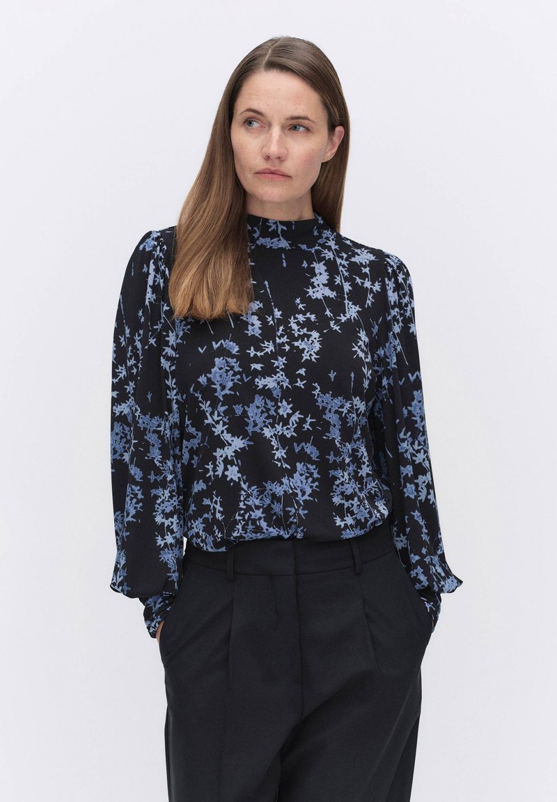 Svart blus med blå blommönster, hög krage och långa ärmar. Slät tyg med avslappnad passform, kombinerad med mörka byxor.