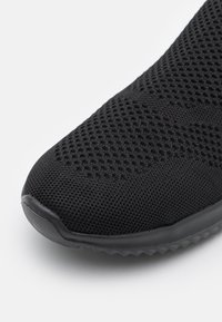 Chaussure de sport noire avec une tige en mesh texturé et respirant et une semelle en caoutchouc. L'avant présente une forme arrondie avec un design minimaliste.