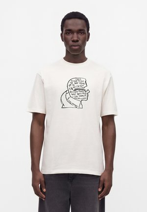 Homme portant un t-shirt blanc avec un dessin au trait noir d'une tête et un texte en profil lisant une citation de Karl Lagerfeld.