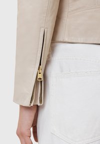 Manica di giacca in pelle beige con una cerniera elegante, dotata di accessori in oro e denti neri in contrasto, finitura testurizzata e cuciture minime.