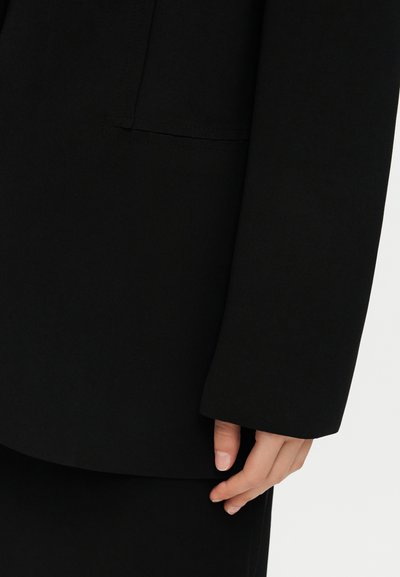 Χέρι ακουμπισμένο δίπλα στο σώμα, φορώντας μαύρο σακάκι με μακριά μανίκια, με ορατή ραφή και λεπτομέρειες τσέπης σε απλό φόντο.