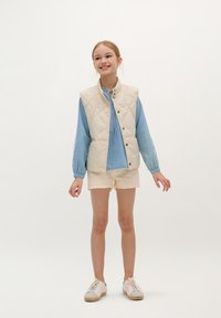 Gilet matelassé crème sur blouse à manches longues bleu clair, associé à un short beige et des baskets blanches. Design simple, coupe décontractée.
