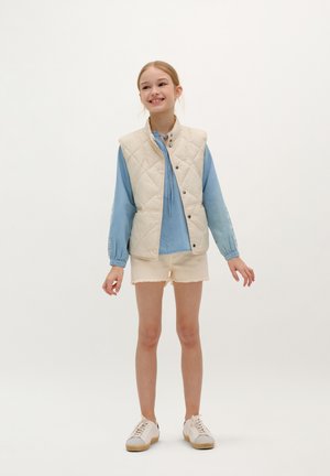 Gilet trapuntato crema sopra una blusa a maniche lunghe azzurro chiaro, abbinato a pantaloni corti beige e sneakers bianche. Design semplice, vestibilità casual.