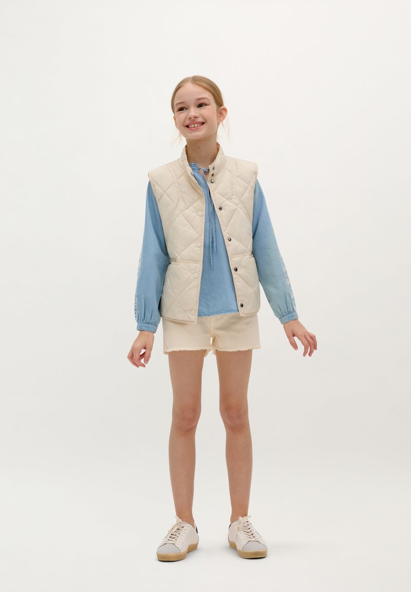 Gilet matelassé crème sur blouse à manches longues bleu clair, associé à un short beige et des baskets blanches. Design simple, coupe décontractée.