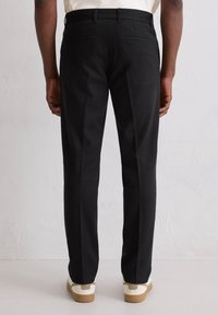 Dopasowane czarne spodnie z gładkim przodem, dwoma tylnymi kieszeniami i wąskim krojem, zestawione z casualowym białym i beżowym obuwiem.