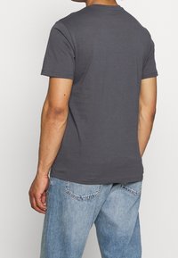 Hombre vestido con una camiseta de manga corta color gris oscuro y unos pantalones de mezclilla azul claro, visto desde atrás sobre un fondo blanco.