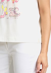 Weißes Baumwoll-T-Shirt mit bunten Schuhillustrationen und dem Text "Verändere dein Leben, frage Cinderella." Kurze Ärmel und lockere Passform.