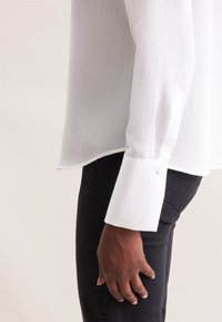 Camicia a righe bianche con maniche lunghe, dotata di polsini con bottone e orlo leggermente curvato. Abbinata a pantaloni di colore scuro.