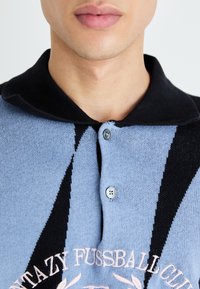 Strikket polo-shirt med et lysblått og sort mønster, to knapper og broderet tekst "FANTAZY FUSSBALL CLUB" i lyserød.