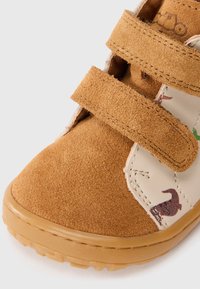 Chaussure en daim marron avec un côté beige arborant des motifs animaliers. Deux bandes Velcro pour la fermeture. Semelle en caoutchouc avec texture antidérapante.