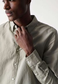 Camisa de botón de un verde claro hecha de tejido suave; presenta un collar clásico, mangas largas y botones blancos en los puños y la tapeta.
