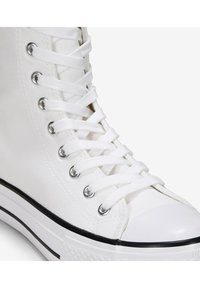 Zapatilla alta blanca con parte superior de lona, punta redonda, suela de goma negra, ojales plateados y cordones blancos enhebrados.