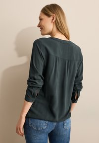 Donkergroene blouse met lange mouwen en een ronde halslijn, geplooide achterkant en met knopen oprolbare mouwen, gecombineerd met blauwe spijkerbroek.