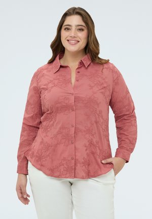 Femme souriante portant une chemise boutonnée à manches longues rose poudré brodée de fleurs et un pantalon blanc, debout avec une main dans la poche.
