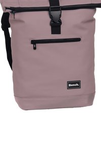 Sac à dos imperméable rose avec des détails de fermeture éclair noire, des bretelles ajustables et une poche zippée à l'avant. Présente une patch de logo en bas.
