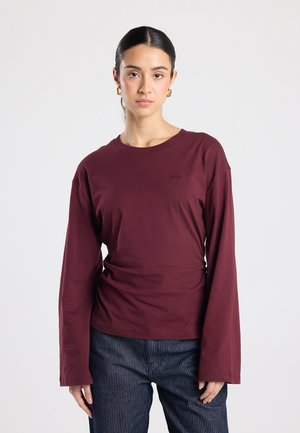 Vrouw met donker haar in een paardenstaart, goudkleurige oorbellen in de vorm van ringen, een lange mouws bordeaux shirt en donkere broek met fijne strepen, staand tegen een effen achtergrond.