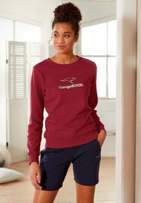 Bordeauxfarbener Sweatshirt mit Rundhalsausschnitt und Känguru-Logo, kombiniert mit dunkelblauen Shorts. Das Material wirkt weich und hat eine legere Passform.