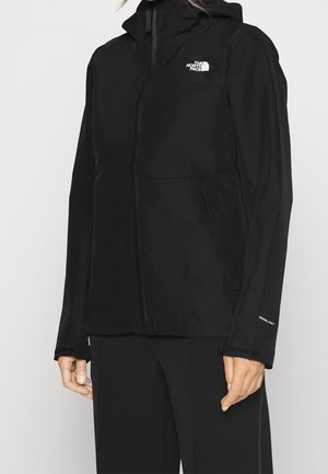 Personne portant une veste noire North Face avec capuche, fermeture éclair à l'avant et poches latérales, associée à un pantalon noir sur un fond uni.