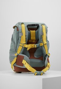 Sac à dos combinant des matériaux verts et bruns, rembourrage en maille, sangles jaunes et accents gris. Conçu pour le confort et le soutien.