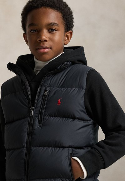 Mladý chlapec nosí černou prošívanou vestu přes černou mikinu s kapucí a bílý rolák, stojí s jednou rukou v kapse před jednobarevným pozadím.