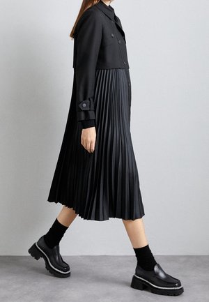 Kvinde går iført en sort cropped jakke, sort plisseret midi-nederdel, sorte sokker og chunky sorte platformsko mod en grå baggrund.