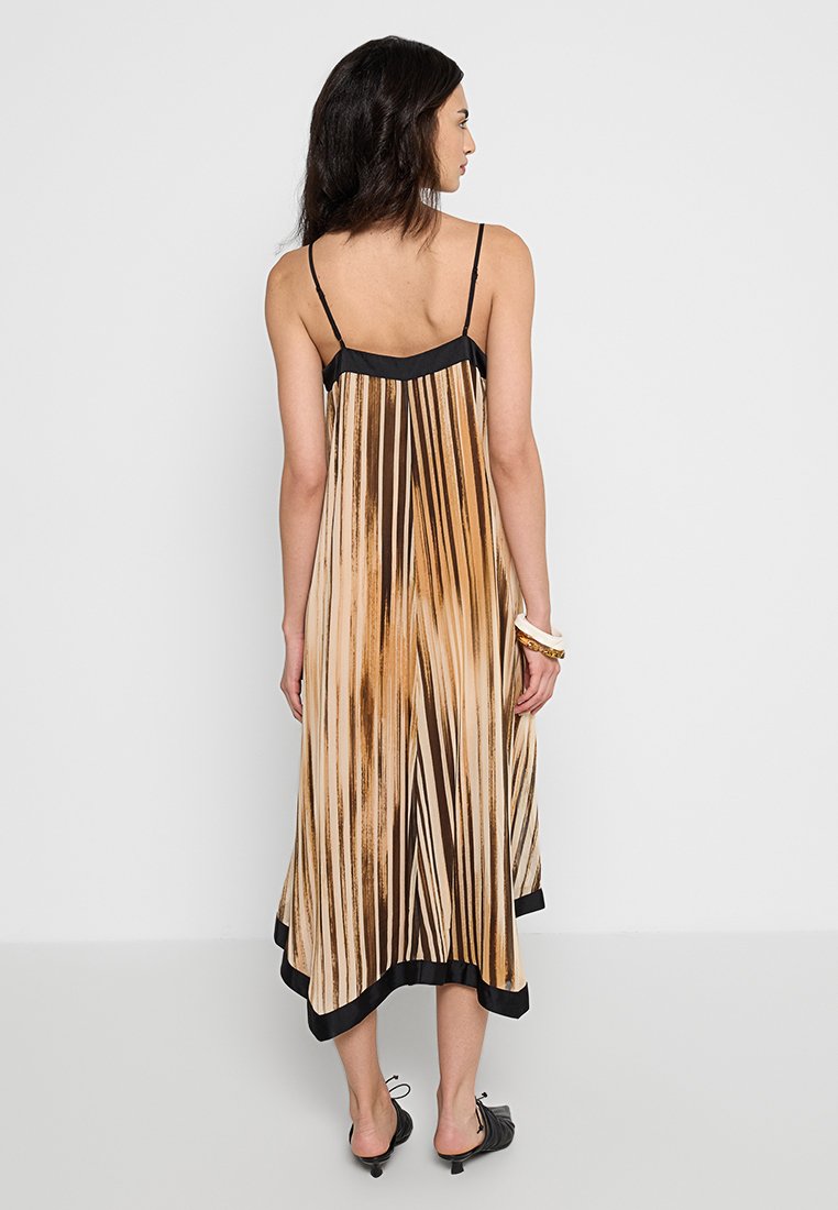 Kobieta w bezrękawnej midi sukience z plisowanym dołem, z brązowymi i beżowymi paskami przypominającymi pociągnięcia pędzla, czarnym wykończeniem, czarnych szpilkach i białych bransoletkach.