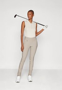 Woman wearing sleeveless beige golf shirt, beige pants, white glove and shoes, holding golf club over shoulder against plain background.