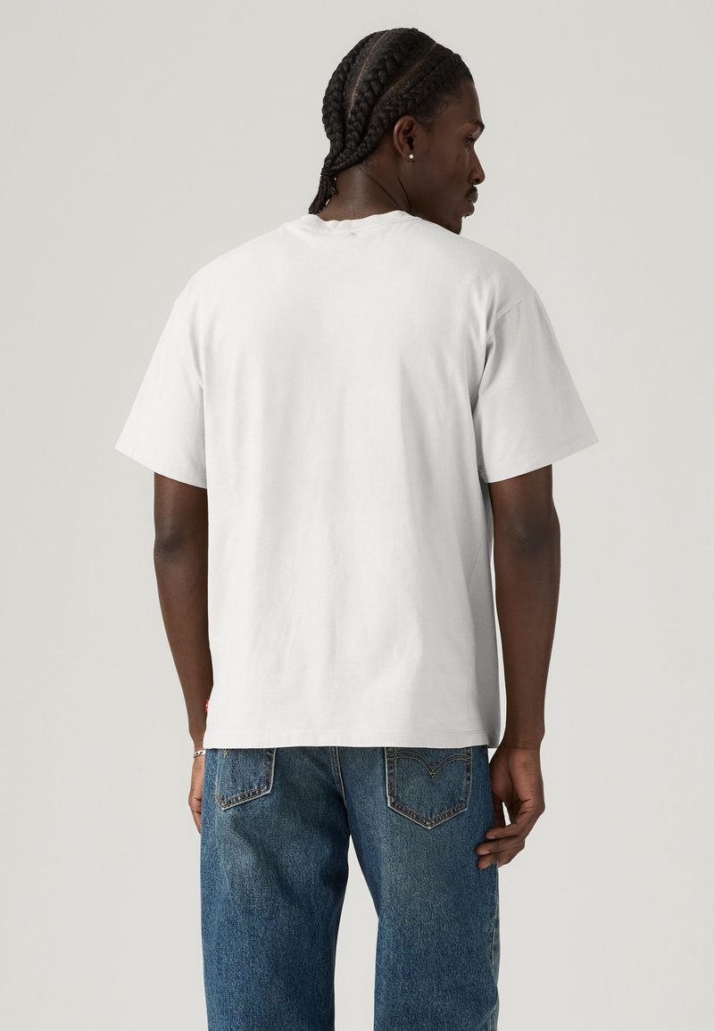 Mand med fletninger iført en simpel hvid T-shirt og blå jeans, set bagfra mod en neutral baggrund.