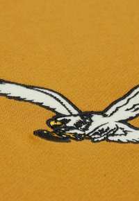 Gelbes Baumwollgewebe mit einem detaillierten schwarz-weißen gestickten Design eines fliegenden Vogels, das die Textur derStickerei und lebendige Farben zeigt.