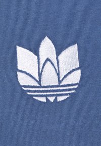 Blått tyg med en texturerad yta som har en vit broderad logotyp som liknar en stiliserad blomma med horisontella linjer under.