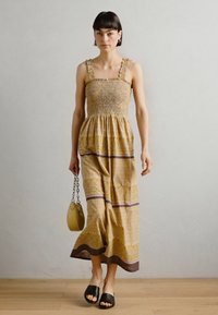 Žuta haljina s uzorkom, smock bodice i naboranim naramenicama. Duga, lepršava konstrukcija s smešnim akcentima i teksturiranim materijalom. Crne cipele na slajd.