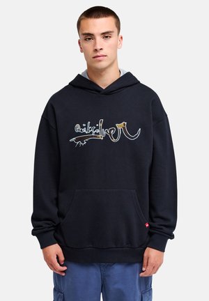 Junger Mann mit schwarzem Hoodie mit grafischem Logo und blauen Cargo-Hosen steht vorwärtsgewandt vor einem schlichten, hellen Hintergrund.