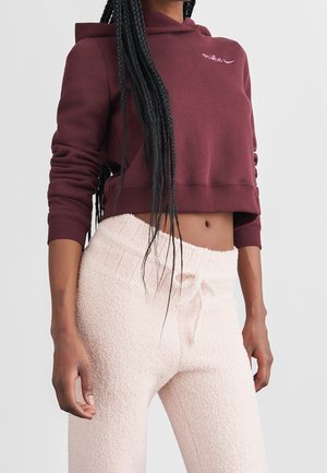 Person trägt einen cropped dunkelroten Nike-Hoodie und hoch taillierte hellrosa strukturierte Hosen mit Kordelzug in der Taille.