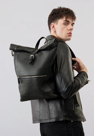 Mladý muž v černé kožené bundě nese velký černý kožený batoh s předním zipem přes rameno a dívá se stranou.