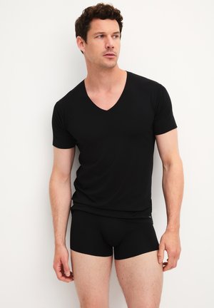Man wearing black V-neck short-sleeve shirt and black boxer briefs standing against white background looking slightly to the side.