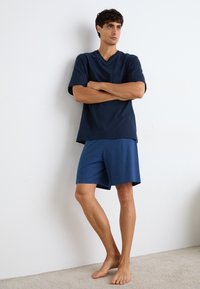 Granatowy t-shirt z dekoltem w serek z bawełny, z kieszonką na piersi, zestawiony z niebieskimi szortami w wzory w romby. Model stoi bosymi stopami na dywanie.