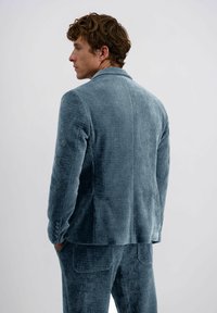 Man met krullend haar, gekleed in een blauwe corduroy pak, zijwaarts geconfronteerd met de handen in de zakken, tegen een effen lichte achtergrond.