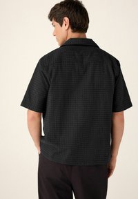 Black short-sleeve shirt with a textured, honeycomb pattern, featuring a notched collar and relaxed fit. Worn by a person seen from the back.