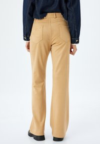 Pantalones acampanados de color marrón claros, hechos de una tela suave, que presentan dos bolsillos traseros con costuras decorativas y un diseño de cintura alta.