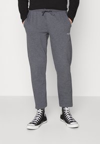 Graue Jogginghosen mit einem Kordelzugbund, tapered Beinen und Seitentaschen. Kombiniert mit schwarzen High-Top-Sneakern. Einfaches Logodesign vorhanden.